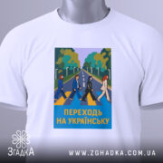 Футболка переходь на українську, біла, зображена в горизонтальному ракурсі, фон світло-сірий, принт з п'ятьма персонажами, що переходять дорогу, на фоні зелених дерев, текст "ПЕРЕХОДЬ НА УКРАЇНСЬКУ" у жовтому кольорі, на комірці є етикетка з логотипом.