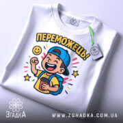 Футболка переможець для дітей. Біла футболка, складена на столі, з яскравим принтом хлопчика у кепці, що тримає чашку. Принт оточений зірочками та написом 'ПЕРЕМОЖЕШЬ!'. На футболці є етикетка з зеленим шнурком.