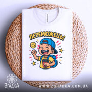 Футболка переможець для дітей. Біла футболка, складена на плетеній основі, з яскравим принтом хлопчика у кепці, що тримає чашку. Принт оточений зірочками та написом 'ПЕРЕМОЖЕШЬ!'. Фон натуральний, що додає затишку.