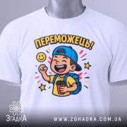 Футболка переможець для дітей. Біла футболка, складена на столі, з яскравим принтом хлопчика у кепці, що тримає чашку. Принт оточений зірочками та написом 'ПЕРЕМОЖЕШЬ!'. На футболці є етикетка з зеленим шнурком.