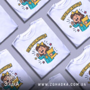 Футболка переможниця білого кольору, зображена на манекені, з принтом, що зображує дівчинку з медаллю та подушкою, фон світло-сірий, деталі принта яскраві та кольорові.