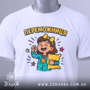 Футболка переможниця білого кольору, складена на поверхні, з принтом дівчинки з медаллю та подушкою, фон світло-сірий, на етикетці зелена стрічка.