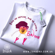 Футболка персик курага багіня, біла футболка складена на поверхні, принт з жовтим персиком у рожевих окулярах, фон світло-сірий, на футболці є етикетка.