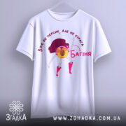 Футболка персик курага багіня, біла футболка підвішена на вішалці, принт з жовтим персиком у рожевих чоботах, фон світло-сірий, текст червоним кольором.