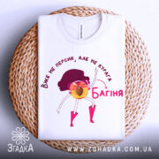 Футболка персик курага багіня, біла футболка на плетеній основі, принт з жовтим персиком у рожевих чоботах, фон світло-сірий, текст червоним кольором.