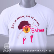 Футболка персик курага багіня, біла футболка складена на поверхні, принт з жовтим персиком у рожевих окулярах, фон світло-сірий, на футболці є етикетка.