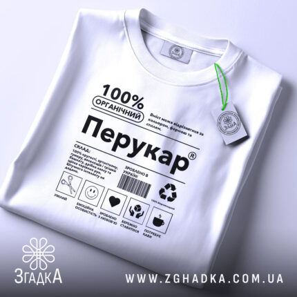 Футболка перукар білого кольору, складена на поверхні, фон світло-сірий, принт чорного кольору з написом '100% ОРГАНІЧНИЙ Перукар' та етикетка з зеленим шнурком.