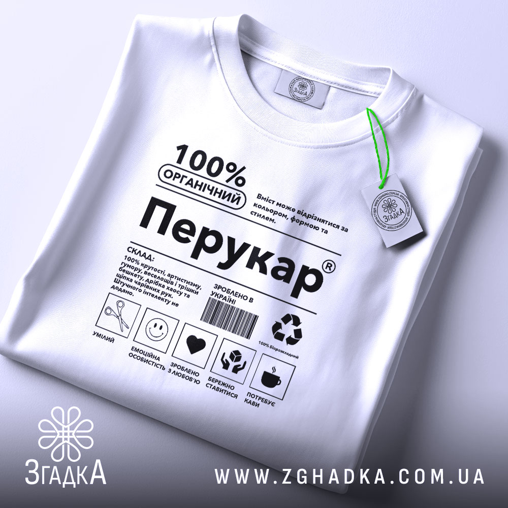 Футболка перукар білого кольору, складена на поверхні, фон світло-сірий, принт чорного кольору з написом '100% ОРГАНІЧНИЙ Перукар' та етикетка з зеленим шнурком.