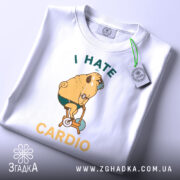 Футболка пес на кардіо, біла, складена на поверхні, фон світло-сірий, принт з жовтим текстом "I HATE CARDIO" та малюнком собаки на велотренажері, з етикеткою зеленого кольору.
