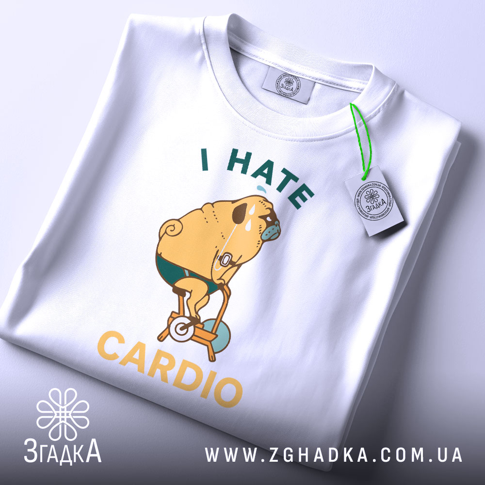 Футболка пес на кардіо, біла, складена на поверхні, фон світло-сірий, принт з жовтим текстом "I HATE CARDIO" та малюнком собаки на велотренажері, з етикеткою зеленого кольору.