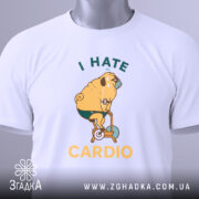 Футболка пес на кардіо, біла, складена на поверхні, фон світло-сірий, принт з жовтим текстом "I HATE CARDIO" та малюнком собаки на велотренажері, з етикеткою зеленого кольору.