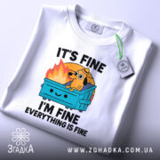 Футболка песик все добре. Біла футболка, складена на столі, з ракурсу зверху. Фон світлий, на футболці яскравий принт з собакою на сміттєвому контейнері, текст "IT'S FINE I'M FINE EVERYTHING IS FINE". На футболці є етикетка з зеленим шнурком.