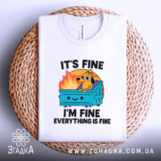 Футболка песик все добре. Біла футболка, складена на плетеній кошику, з ракурсу зверху. Фон світлий, принт яскравий, з собакою на сміттєвому контейнері, текст "IT'S FINE I'M FINE EVERYTHING IS FINE".