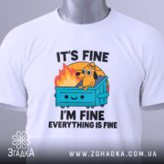 Футболка песик все добре. Біла футболка, складена на столі, з ракурсу зверху. Фон світлий, на футболці яскравий принт з собакою на сміттєвому контейнері, текст "IT'S FINE I'M FINE EVERYTHING IS FINE". На футболці є етикетка з зеленим шнурком.