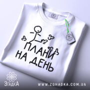 Футболка плани на день, білого кольору, зображена зверху, складена на білому фоні, принт з малюнком stickman і написом "ПЛАНИ НА ДЕНЬ" чорного кольору, на комірці видно етикетку з зеленим шнурком.