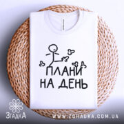 Футболка плани на день, білого кольору, складена на плетеній кошику, фон світло-сірий, принт з малюнком stickman і написом "ПЛАНИ НА ДЕНЬ" чорного кольору, видно округлий комір.