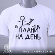 Футболка плани на день, білого кольору, зображена зверху, складена на білому фоні, принт з малюнком stickman і написом "ПЛАНИ НА ДЕНЬ" чорного кольору, на комірці видно етикетку з зеленим шнурком.