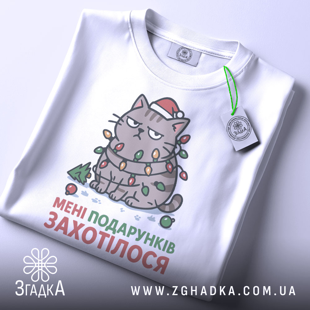 Футболка подарунків захотілося, біла, зображена з верхнього ракурсу, на фоні світло-сірого кольору, принт з котом в червоній шапці, обмотаним гірляндою, з текстом "МЕНІ ПОДАРУНКІВ ЗАХОТІЛОСЯ", на футболці є етикетка з зеленим шнурком.