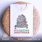 Футболка подарунків захотілося, біла, зображена зверху, на фоні плетеного кошика, принт з котом в червоній шапці, обмотаним гірляндою, з текстом "МЕНІ ПОДАРУНКІВ ЗАХОТІЛОСЯ", складена на плетеній основі.