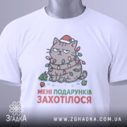 Футболка подарунків захотілося, біла, зображена з верхнього ракурсу, на фоні світло-сірого кольору, принт з котом в червоній шапці, обмотаним гірляндою, з текстом "МЕНІ ПОДАРУНКІВ ЗАХОТІЛОСЯ", на футболці є етикетка з зеленим шнурком.