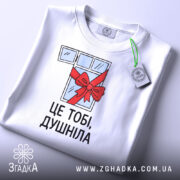 Футболка подарунок душнілі, біла, зображена зверху, на ній видно етикетку з зеленим шнурком, фон світло-сірий, принт з малюнком вікна з червоною стрічкою та написом 'ЦЕ ТОБІ, ДУШНІЛА'.
