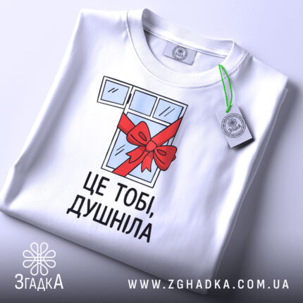 Футболка подарунок душнілі, біла, зображена зверху, на ній видно етикетку з зеленим шнурком, фон світло-сірий, принт з малюнком вікна з червоною стрічкою та написом 'ЦЕ ТОБІ, ДУШНІЛА'.