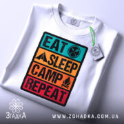 Футболка подорожуй легко, біла, складена на столі, фон світло-сірий, принт з написом 'EAT SLEEP CAMP REPEAT' у яскравих кольорах, з етикеткою на комірці.