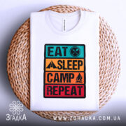 Футболка подорожуй легко, біла, складена на плетеному кошику, фон світло-сірий, принт з написом 'EAT SLEEP CAMP REPEAT' у яскравих кольорах, видно деталі тканини та принта.