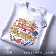 Футболка покликання вчителя білого кольору, складена на поверхні, фон світлий, принт з текстом "ВЧИТЕЛЬ НЕ РОБОТА, А ПОКЛИКАННЯ" та малюнками олівців і яблука, на футболці є етикетка.