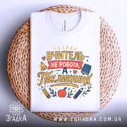 Футболка покликання вчителя білого кольору, складена на круглій плетеній основі, фон нейтральний, принт з текстом "ВЧИТЕЛЬ НЕ РОБОТА, А ПОКЛИКАННЯ" та малюнками олівців і яблука.