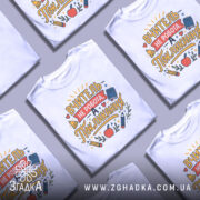 Футболка покликання вчителя білого кольору, зображена з близького ракурсу, фон нейтральний, принт містить текст "ВЧИТЕЛЬ НЕ РОБОТА, А ПОКЛИКАННЯ" з ілюстраціями олівців, яблука та книжки.