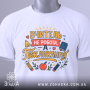 Футболка покликання вчителя білого кольору, складена на поверхні, фон світлий, принт з текстом "ВЧИТЕЛЬ НЕ РОБОТА, А ПОКЛИКАННЯ" та малюнками олівців і яблука, на футболці є етикетка.