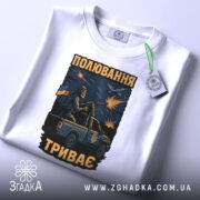 Футболка полювання триває, біла футболка на столі, ракурс зверху, фон світлий, принт з військовим на пікапі, з тегом на зеленій стрічці.