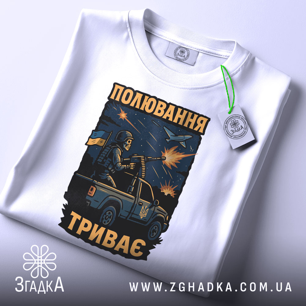 Футболка полювання триває, біла футболка на столі, ракурс зверху, фон світлий, принт з військовим на пікапі, з тегом на зеленій стрічці.