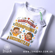 Футболка помічник вихователя білого кольору, зображена зверху, на ній видно етикетку, фон світло-сірий, принт містить напис 'НАЙКРАЩИЙ ПОМІЧНИК ВИХОВАТЕЛЯ' з ілюстраціями дітей, які радісно піднімають руки, оточений кольоровими зірочками та елементами декору.