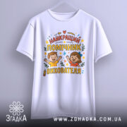 Футболка помічник вихователя білого кольору, зображена з фронтального ракурсу, фон світло-сірий, принт містить напис 'НАЙКРАЩИЙ ПОМІЧНИК ВИХОВАТЕЛЯ' з ілюстраціями дітей, які радісно піднімають руки, оточений кольоровими зірочками та елементами декору.