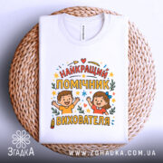 Футболка помічник вихователя білого кольору, зображена зверху на плетеній кошику, фон світло-сірий, принт містить напис 'НАЙКРАЩИЙ ПОМІЧНИК ВИХОВАТЕЛЯ' з ілюстраціями дітей, які радісно піднімають руки, оточений кольоровими зірочками та елементами декору.