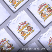 Футболка помічник вихователя білого кольору, зображена з правого ракурсу на манекені, фон світло-сірий, принт містить напис 'НАЙКРАЩИЙ ПОМІЧНИК ВИХОВАТЕЛЯ' з ілюстраціями дітей, які радісно піднімають руки, оточений кольоровими зірочками та елементами декору.