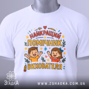 Футболка помічник вихователя білого кольору, зображена зверху, на ній видно етикетку, фон світло-сірий, принт містить напис 'НАЙКРАЩИЙ ПОМІЧНИК ВИХОВАТЕЛЯ' з ілюстраціями дітей, які радісно піднімають руки, оточений кольоровими зірочками та елементами декору.