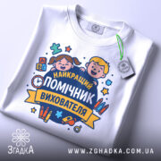 Футболка помічник вихователя, біла, складена на столі, фон світло-сірий, принт з двома усміхненими дітьми та текстом 'НАЙКРАЩИЙ ПОМІЧНИК ВИХОВАТЕЛЯ', на футболці є етикетка.
