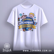 Футболка помічник вихователя, біла, підвішена на вішалці, фон світло-сірий, принт з дітьми та текстом 'НАЙКРАЩИЙ ПОМІЧНИК ВИХОВАТЕЛЯ', видно деталі дизайну.