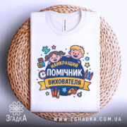 Футболка помічник вихователя, біла, складена на плетеному кошику, фон світлий, принт з усміхненими дітьми та текстом 'НАЙКРАЩИЙ ПОМІЧНИК ВИХОВАТЕЛЯ', виглядає акуратно.