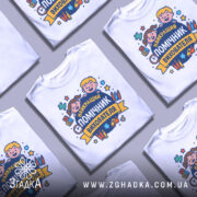 Футболка помічник вихователя, біла, зображена з правого ракурсу на манекені, фон світло-сірий, принт з веселими дітьми та текстом 'НАЙКРАЩИЙ ПОМІЧНИК ВИХОВАТЕЛЯ' у яскравих кольорах.