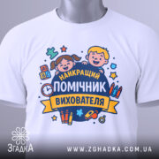 Футболка помічник вихователя, біла, складена на столі, фон світло-сірий, принт з двома усміхненими дітьми та текстом 'НАЙКРАЩИЙ ПОМІЧНИК ВИХОВАТЕЛЯ', на футболці є етикетка.