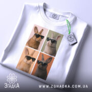 Футболка попарт кролики, біла, складена на поверхні, фон білий, принт з чотирма кроликами в окулярах, два з них коричневі, два світлі, з тегом на зеленому шнурку.