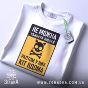 Футболка попередження про кота, біла, складена на поверхні, фон світло-сірий, принт з чорним написом і жовтим фоном, з малюнком кота і кісток, з етикеткою на зеленому шнурку.
