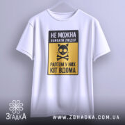 Футболка попередження про кота, біла, підвішена на вішалці, фон світло-сірий, принт з чорним написом і жовтим фоном, з малюнком кота і кісток.