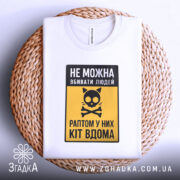 Футболка попередження про кота, біла, складена на плетеній кошику, фон білий, принт з чорним написом і жовтим фоном, з малюнком кота і кісток.