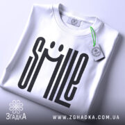 Футболка посміхнись. Біла футболка, складена на столі, фон світло-сірий, принт з написом 'SMILE' з усмішкою, на ярлику зелена стрічка.