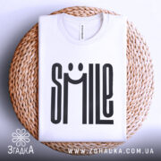 Футболка посміхнись. Біла футболка, складена на плетеній кошику, фон світло-сірий, принт з написом 'SMILE' з усмішкою, видно круглий виріз горловини.